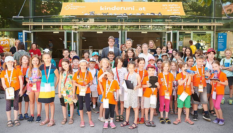 Kinder stehen im Uni Campus vor dem Hörsaal. die meisten tragen ein oranges Shirt. Bildungsminister Martin Polaschek und Vizerektorin Schnabel stehen bei den Kindern.